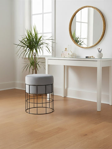 A velvet stool with a gray cushion and a sleek metal cage frame sits on a light wooden floor next to a white console table. A round mirror with a gold frame hangs on the wall above the table, and a potted plant adds a touch of greenery to the scene. Natural light streams in through a window, creating a bright and inviting atmosphere