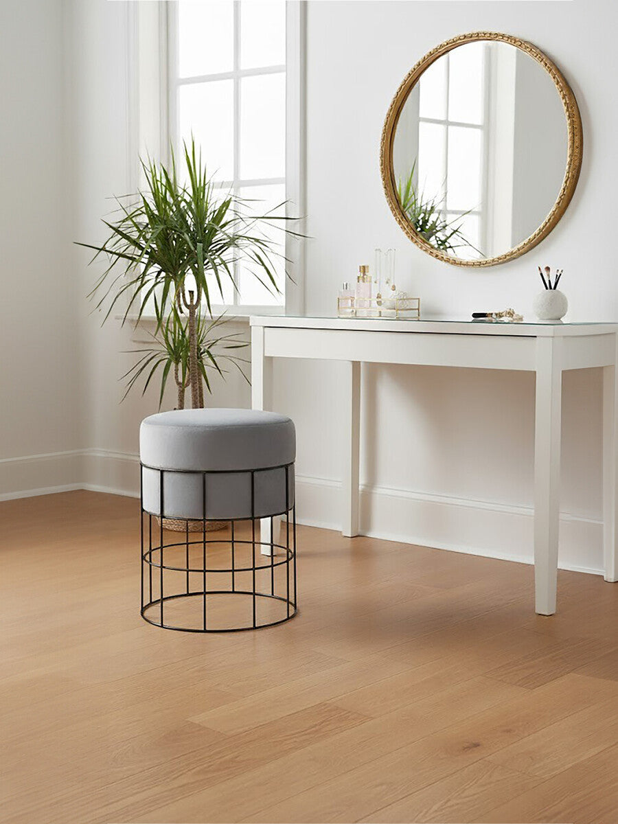 A velvet stool with a gray cushion and a sleek metal cage frame sits on a light wooden floor next to a white console table. A round mirror with a gold frame hangs on the wall above the table, and a potted plant adds a touch of greenery to the scene. Natural light streams in through a window, creating a bright and inviting atmosphere