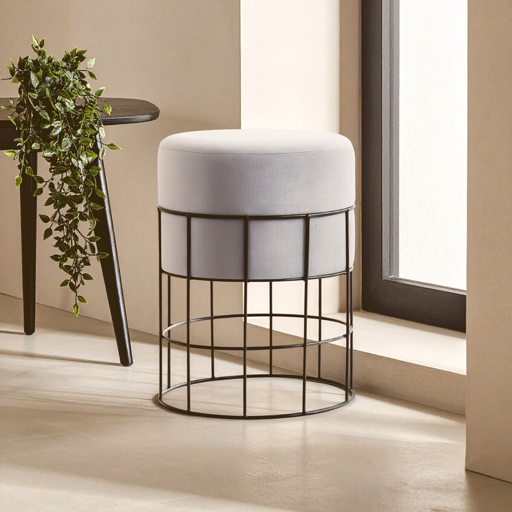 A velvet stool with a round, cushioned top in light gray, supported by a black metal cage frame. The stool is positioned near a window with natural light illuminating the scene, and a small potted plant is visible on a nearby table