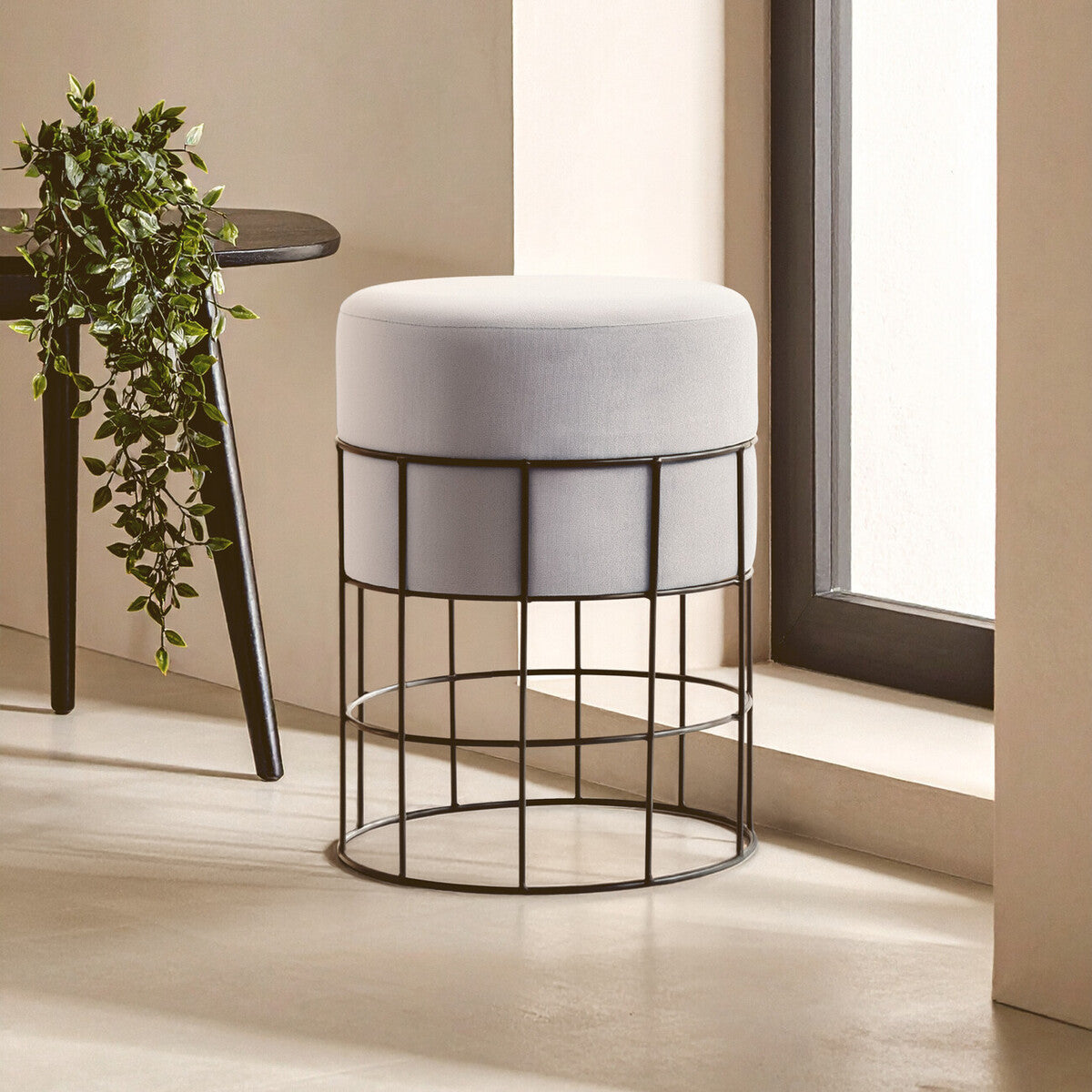 A velvet stool with a round, cushioned top in light gray, supported by a black metal cage frame. The stool is positioned near a window with natural light illuminating the scene, and a small potted plant is visible on a nearby table