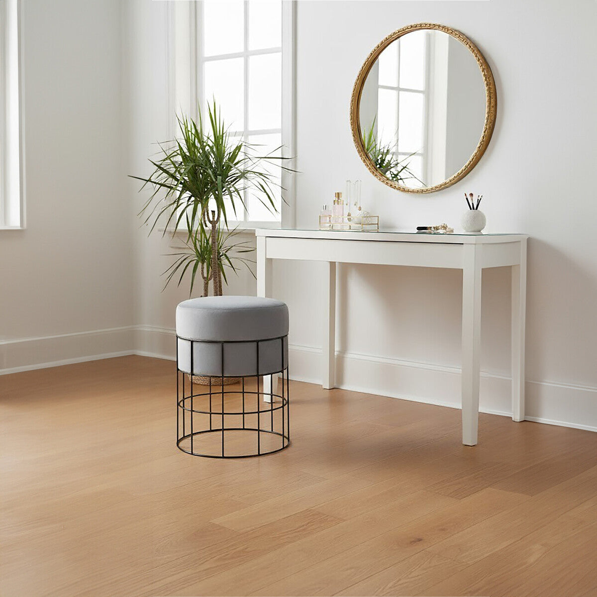 A velvet stool with a gray top and a gold cage frame sits on a light wooden floor in a bright room. A round mirror hangs above a white console table, and a potted plant is positioned nearby, adding a touch of greenery to the space. Natural light streams in through large windows, illuminating the scene