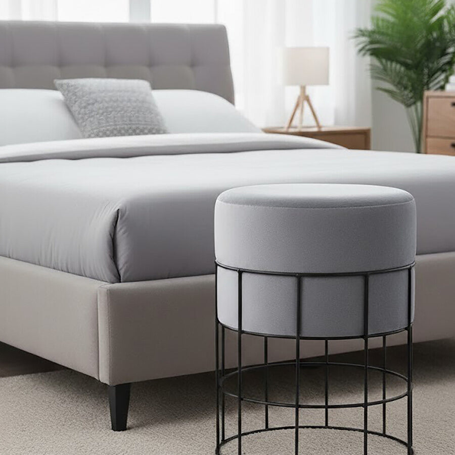 A round velvet stool in light gray sits beside a bed with a gray upholstered headboard. The stool features a black metal cage frame, and the room has a neutral-colored carpet and a bedside lamp in the background. A potted plant adds a touch of greenery to the space