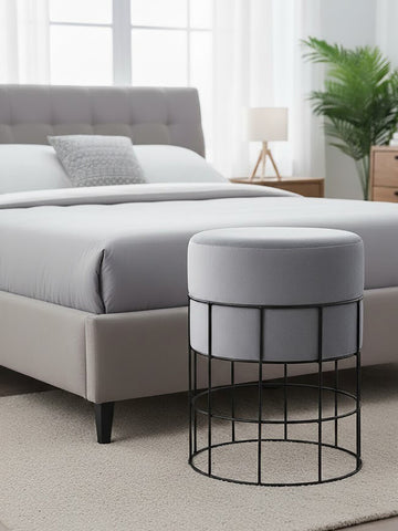 A round velvet stool in light gray sits beside a bed with a gray upholstered headboard. The stool features a black metal cage frame, and the room has a neutral-colored carpet and a bedside lamp in the background. A potted plant adds a touch of greenery to the space