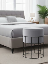 A round velvet stool in light gray sits beside a bed with a gray upholstered headboard. The stool features a black metal cage frame, and the room has a neutral-colored carpet and a bedside lamp in the background. A potted plant adds a touch of greenery to the space