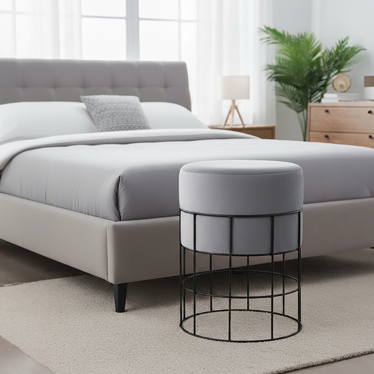 A velvet stool with a round top in light gray, supported by a black metal cage frame, positioned next to a bed with a gray upholstered headboard. The room features a neutral-colored rug, a bedside table with a lamp, and a potted plant in the background