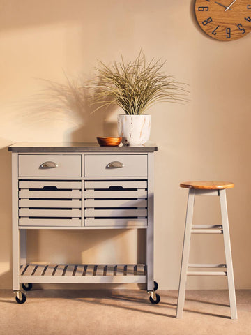 A natural and grey wood bar or kitchen stool with a simple design, featuring a light wood seat and a whitewashed frame. The stool is positioned next to a kitchen cart with two drawers, a decorative plant, and a clock on the wall in a warm, inviting room