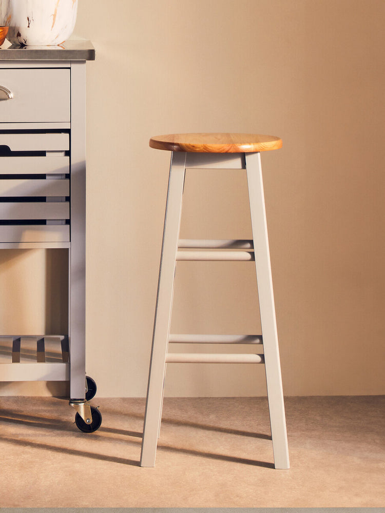 Natural wood seat with a light grey frame bar or kitchen stool, positioned next to a kitchen cart in a softly lit room. The stool features a simple, modern design with four legs and a sturdy construction