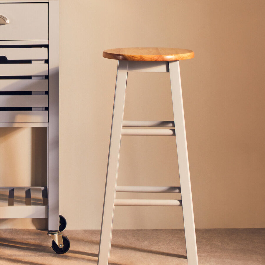 Natural wood seat with a light grey frame bar or kitchen stool, positioned next to a kitchen cart in a softly lit room. The stool features a simple, modern design with four legs and a sturdy construction