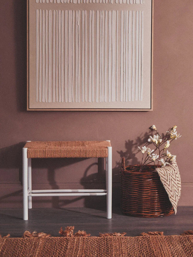 Woven seat wooden bench with white frame beside wicker basket and neutral wall art