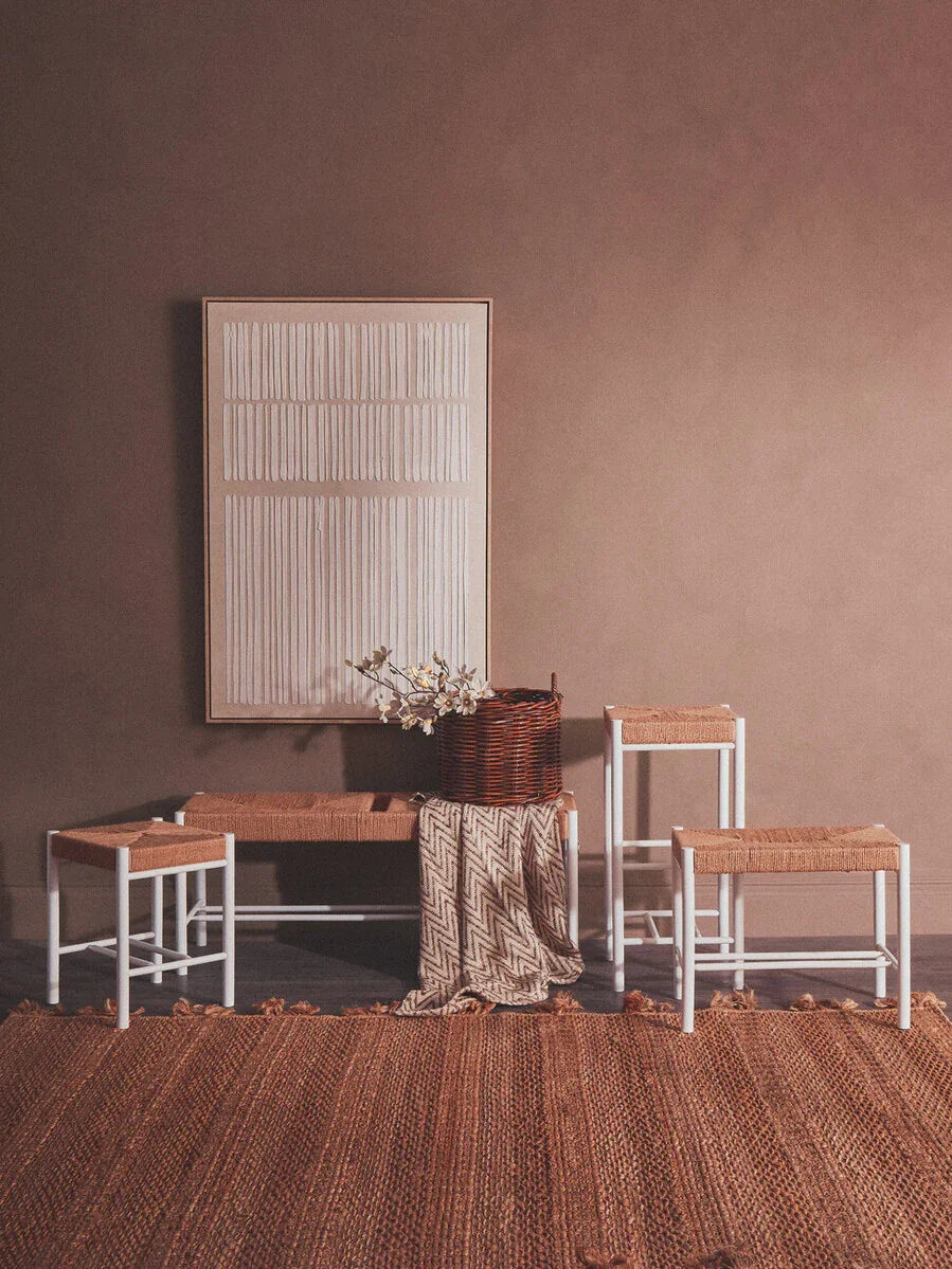 Woven seat stools and bench with white frames, styled on a textured brown rug and wall art