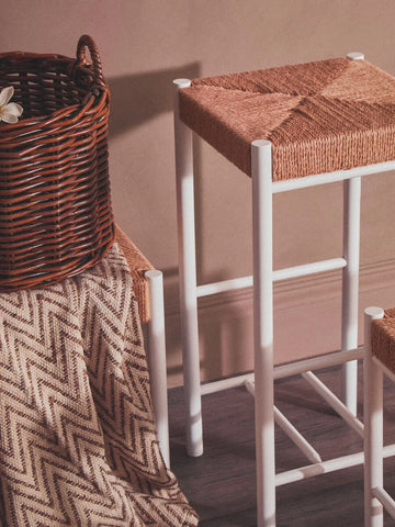 White bar stool with woven rattan seat beside brown wicker basket and chevron throw blanket