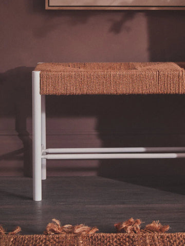 Woven bench with natural fibre seat and white metal legs against dark wall and floor