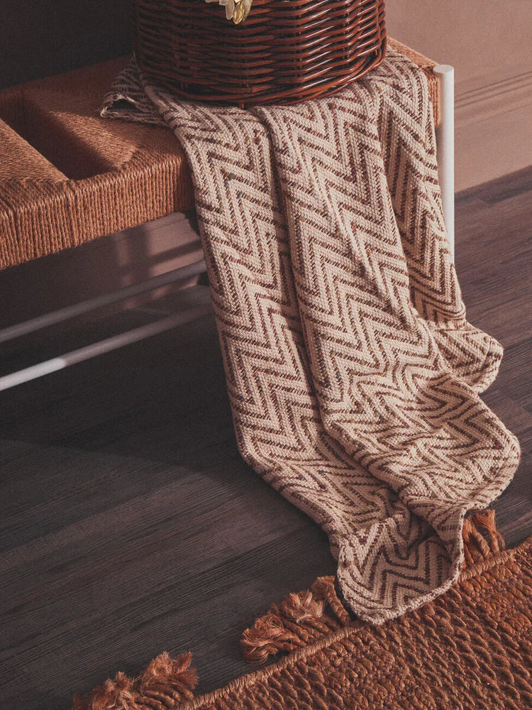 Chevron patterned throw blanket draped over woven bench with tasselled edge on dark wood floor