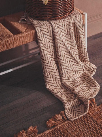 Chevron patterned throw blanket draped over woven bench with tasselled edge on dark wood floor