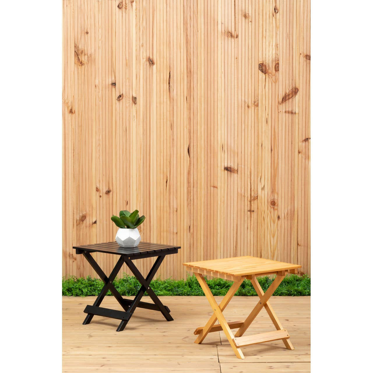 Black wood folding table positioned next to a lighter wood folding table, both featuring an X-shaped base. A small potted plant sits on top of the black table. The background consists of a wooden fence, creating a natural outdoor setting