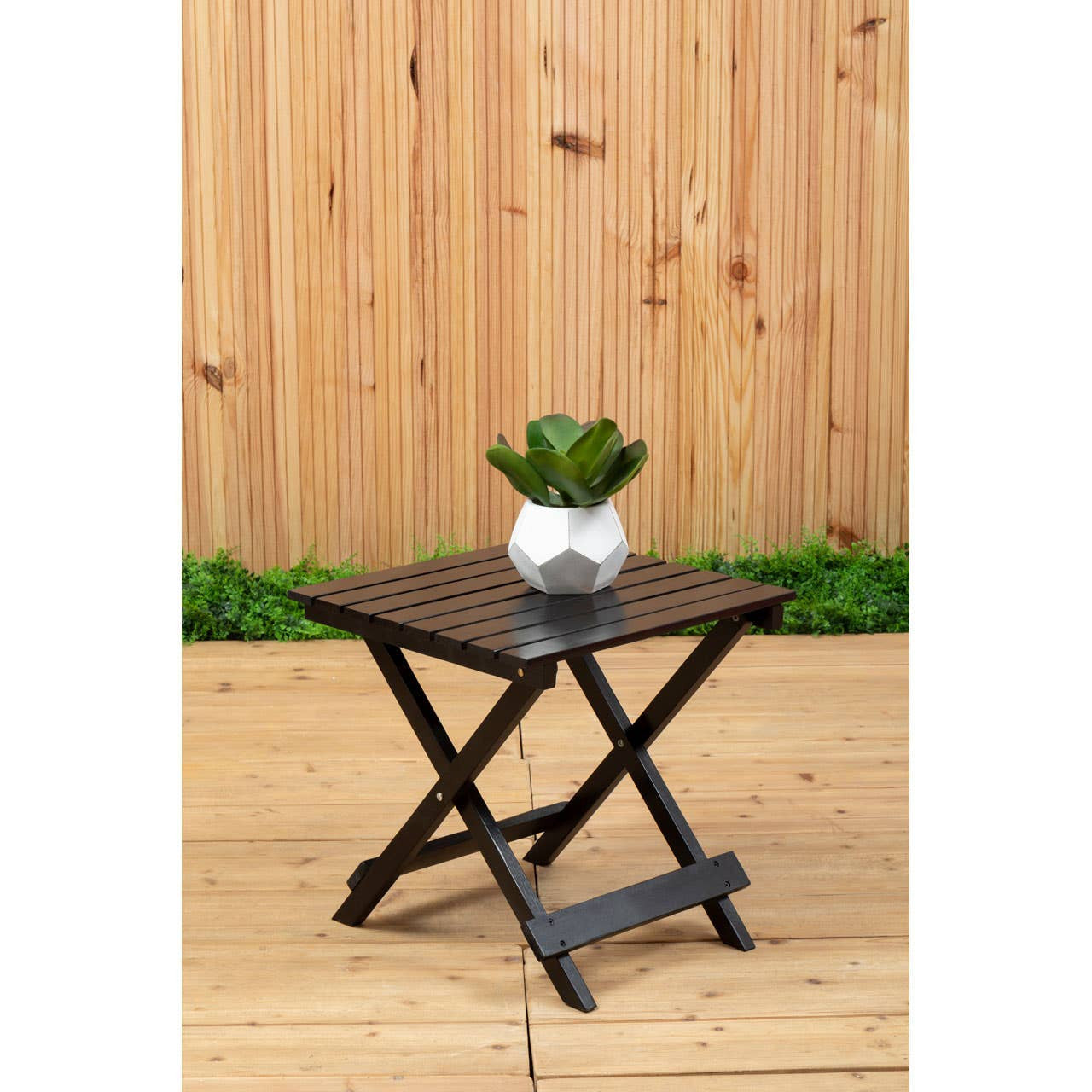 Black wood folding table with a square top, featuring crossed legs for support. A small potted plant sits on the table, surrounded by a wooden deck and a wooden fence in the background