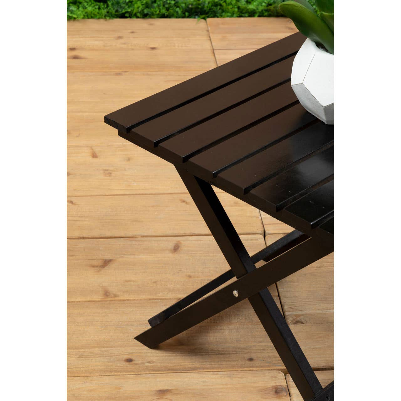 Black wood folding table with slatted top and sturdy crossed legs, placed on a wooden floor with green foliage in the background