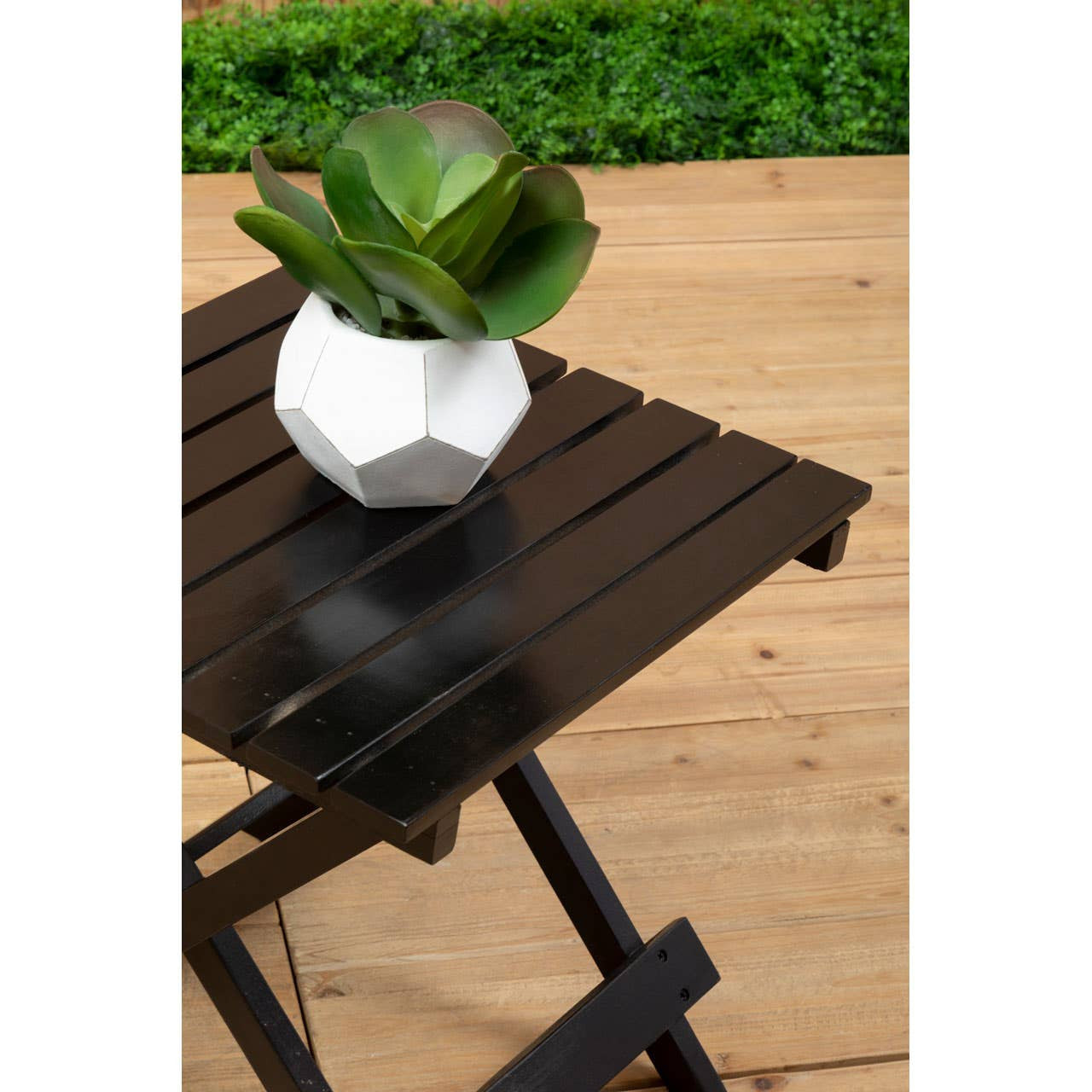 Black wood folding table with a slatted top, featuring a geometric white planter containing a green succulent, set on a wooden surface with grass in the background