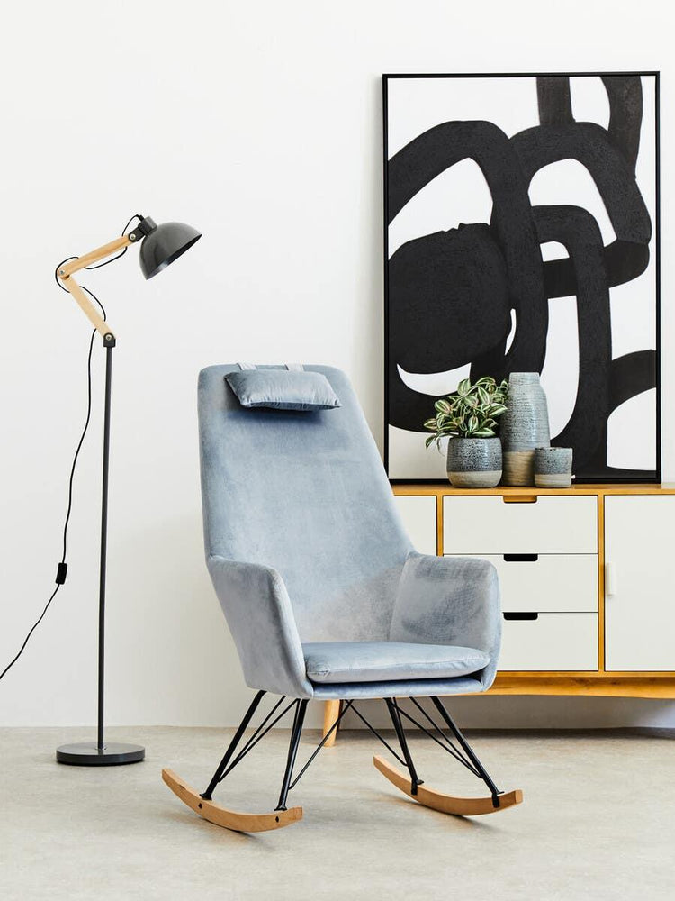 Modern grey velvet rocking chair with metal legs beside black floor lamp and sideboard