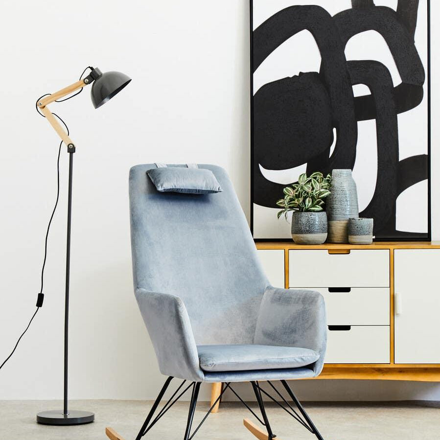 Modern grey velvet rocking chair with metal legs beside black floor lamp and sideboard