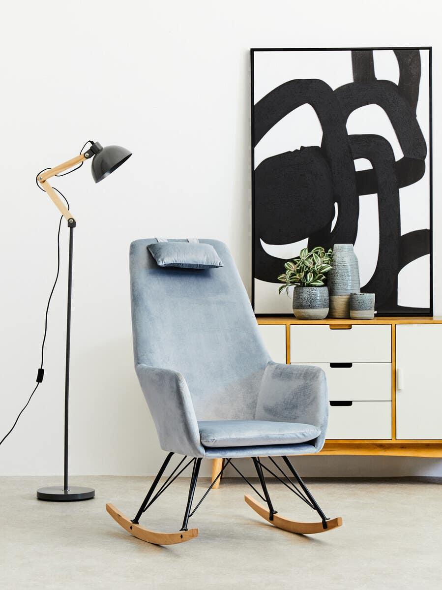 Modern grey velvet rocking chair with metal legs beside black floor lamp and sideboard