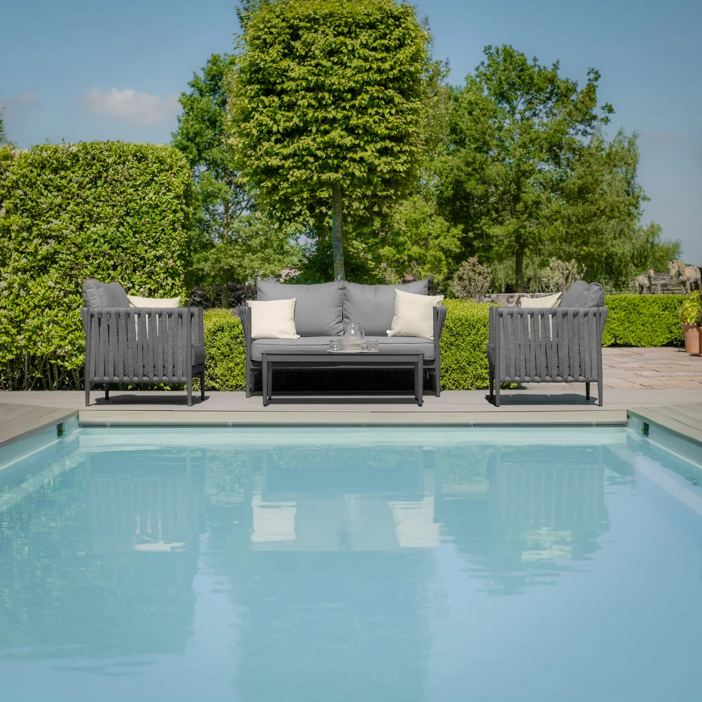 Grey outdoor sofa set with cushions and coffee table beside pool, surrounded by greenery