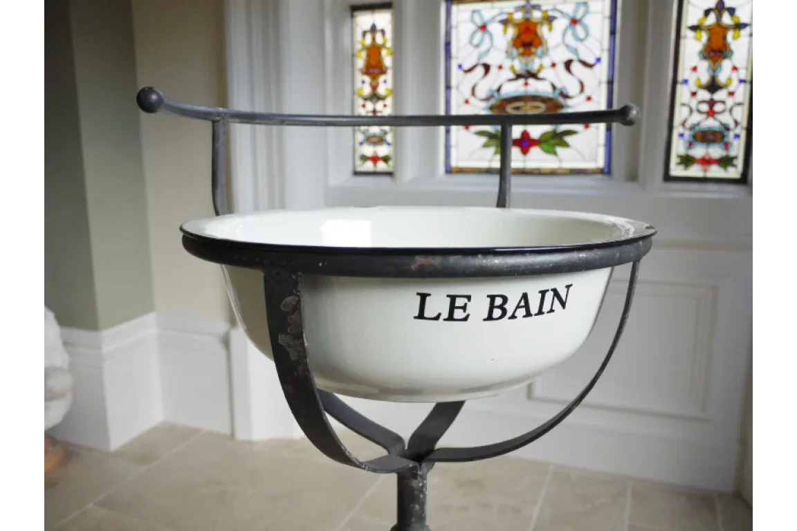 Vintage enamel wash basin on black metal stand with 'Le Bain' text, stained glass background