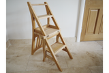 Unfinished light wood chair with foldable steps, shown at an angle on a beige tiled floor near a wooden door.