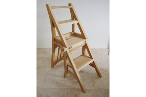 Unfinished light wood folding chair with built-in step ladder, angled view on beige tile floor against white wall