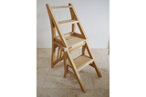 Unfinished light wood folding chair with built-in step ladder, angled view on beige tile floor against white wall