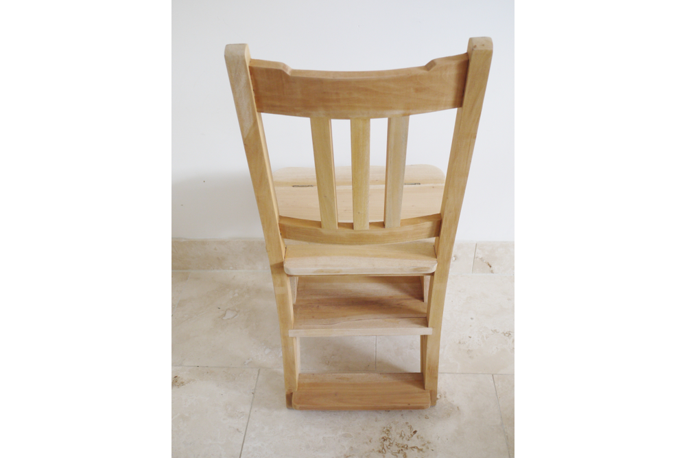 Unfinished wooden step chair with vertical slats, viewed from the back on a tiled floor against a white wall.