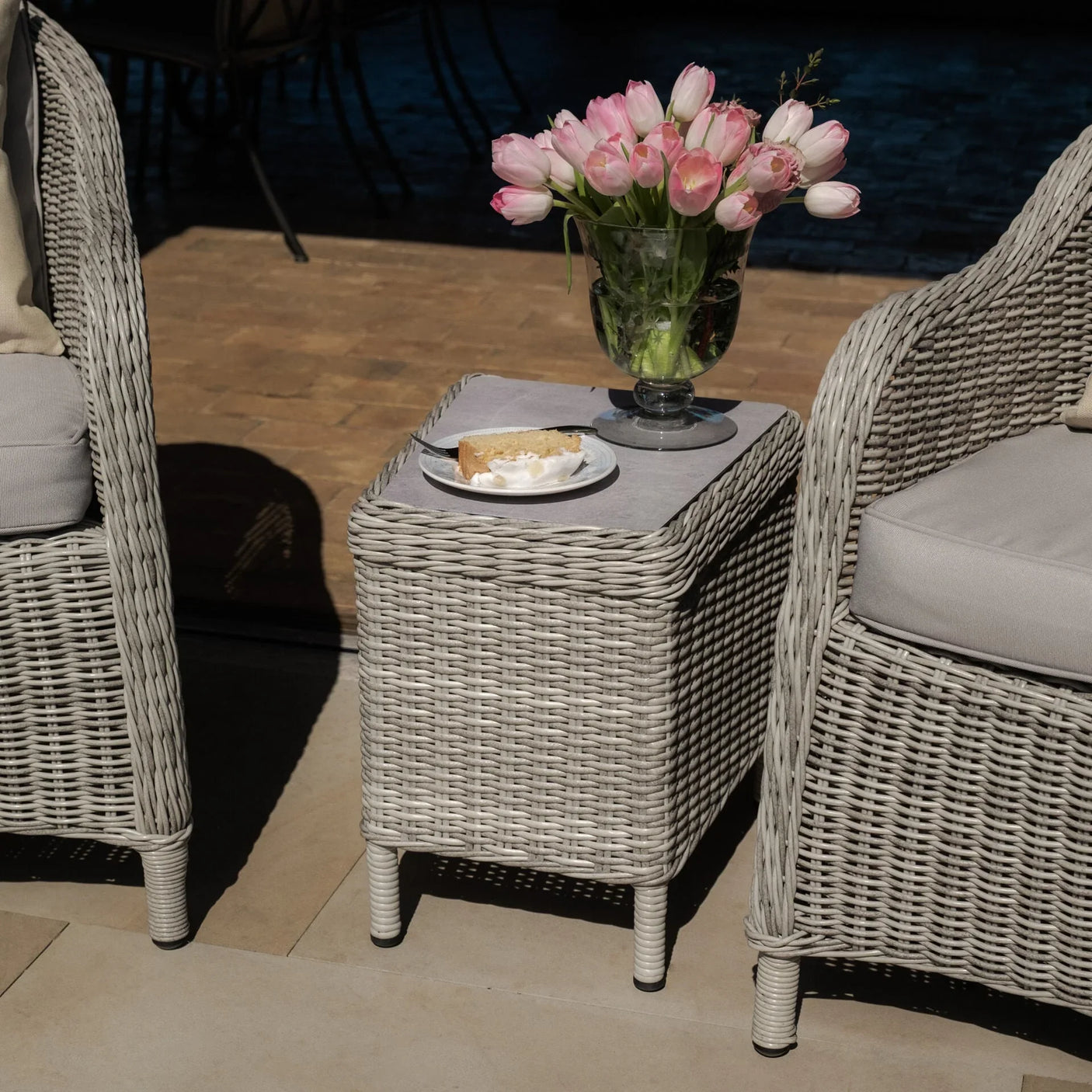 Grey rattan garden side table with glass vase of pink tulips and slice of cake on plate