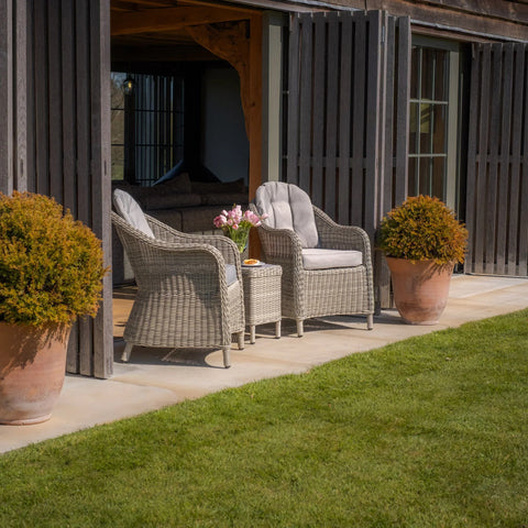 Grey rattan garden chairs with cushions and table on patio beside large potted plants