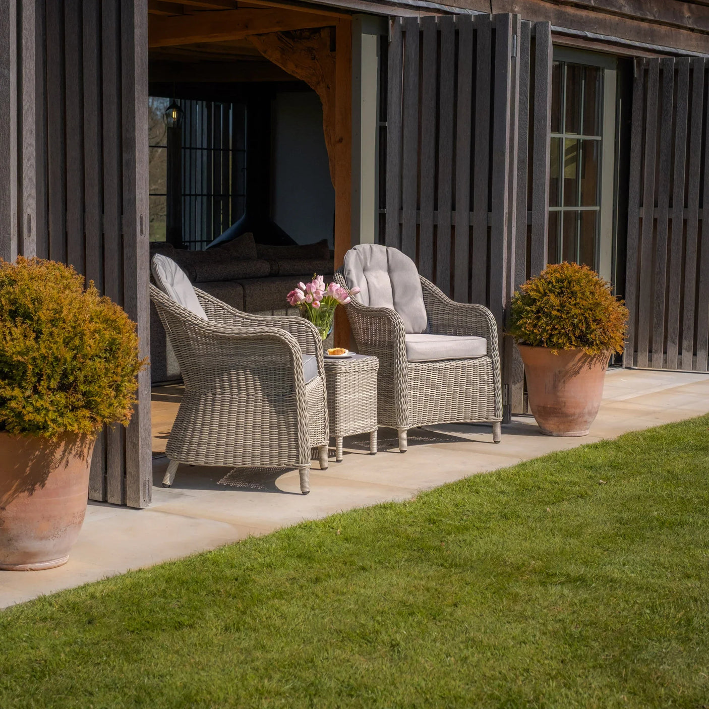 Grey rattan garden chairs with cushions and table on patio beside large potted plants
