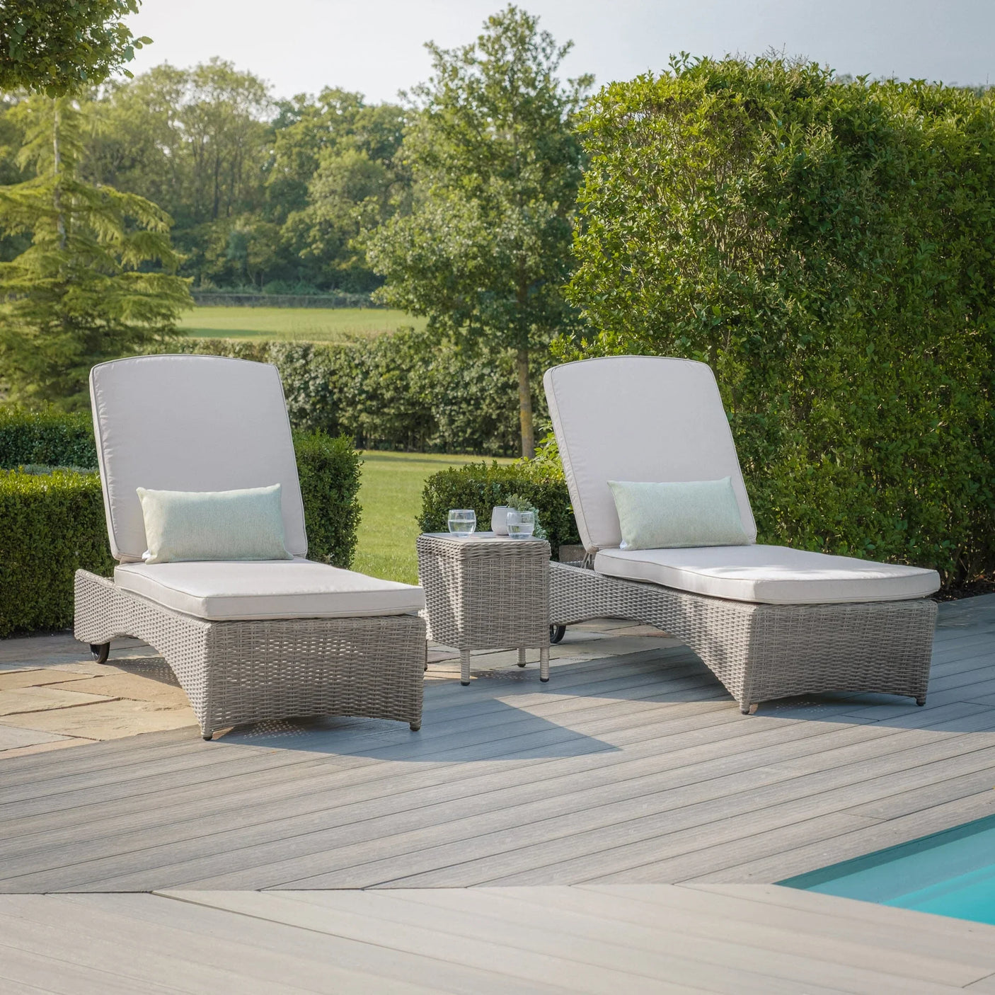 Grey rattan sun loungers with cream cushions and side table by a pool on patio