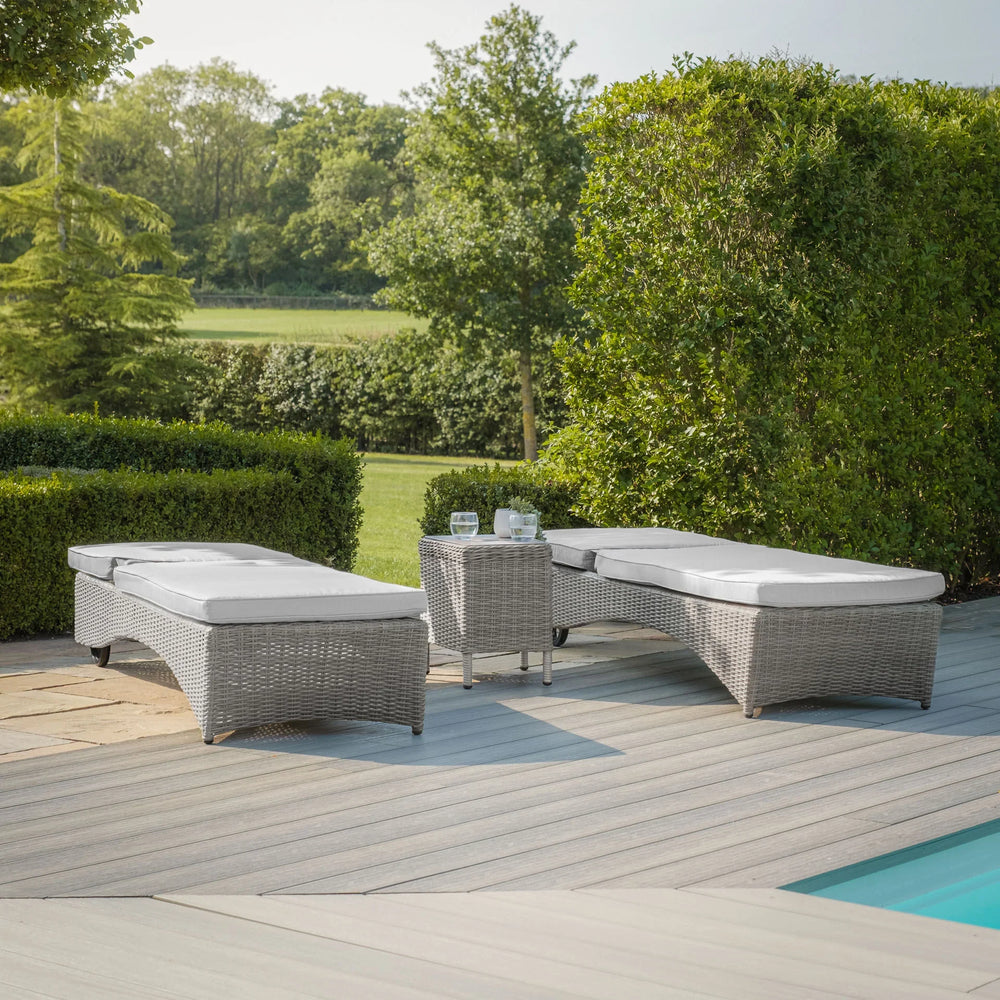 Grey rattan outdoor sun loungers with cushions beside pool on patio with green garden view