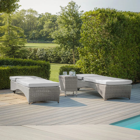 Grey rattan outdoor sun loungers with cushions beside pool on patio with green garden view
