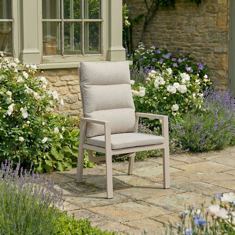 Beige cushioned garden chair on stone patio with flowering shrubs and rustic wall
