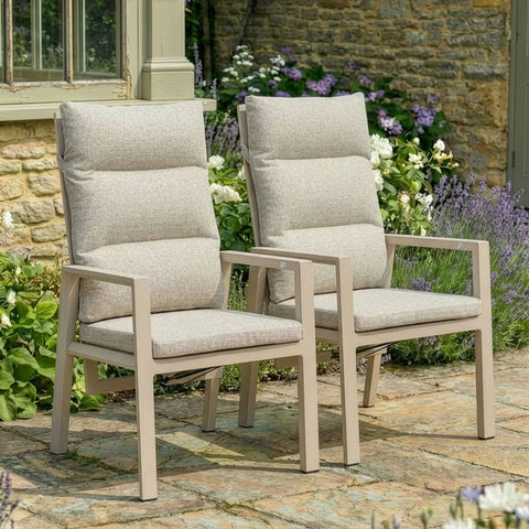 Beige cushioned garden dining chairs on stone patio with flowers and stone wall background