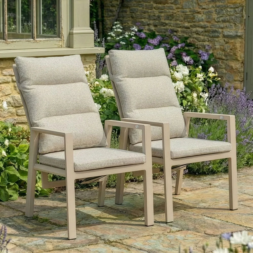 Pair of beige cushioned garden chairs with armrests on stone patio surrounded by flowers