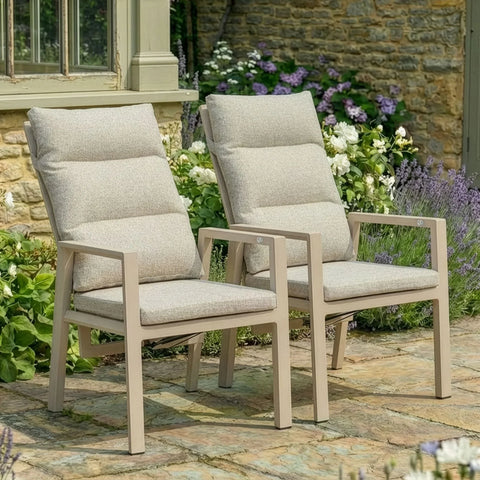 Pair of beige cushioned garden chairs with armrests on stone patio surrounded by flowers