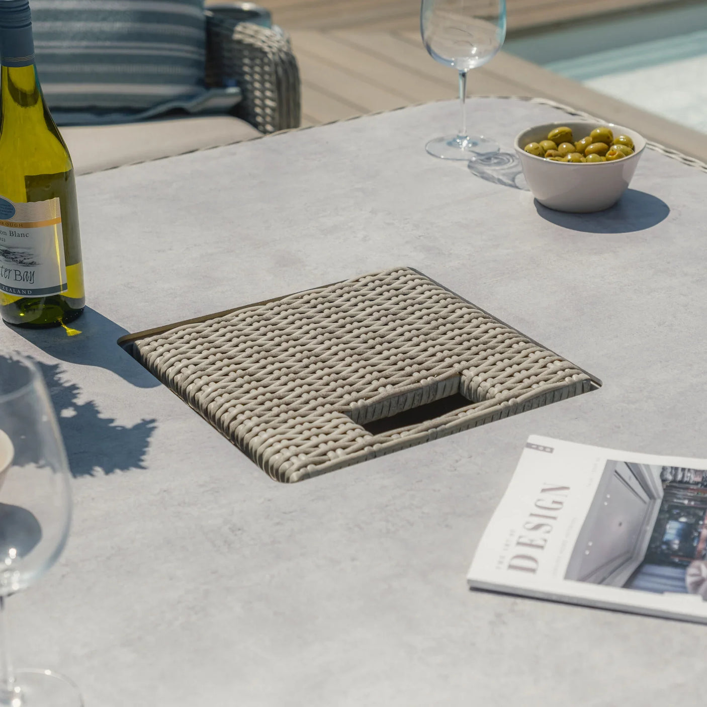 Grey rattan outdoor dining table with wine bottle, glass, bowl of olives and magazine