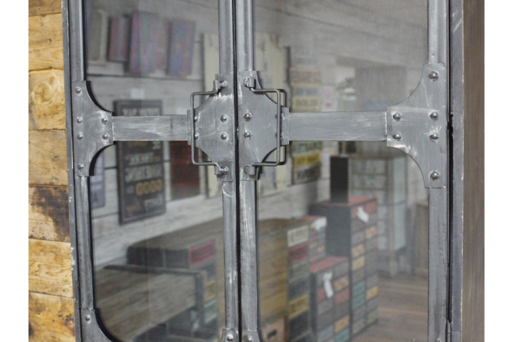 Metal wall cabinet with riveted frame and glass doors, shown in close-up front view in rustic room setting.