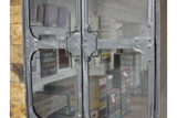 Metal wall cabinet with riveted frame and glass doors, shown in close-up front view in rustic room setting.