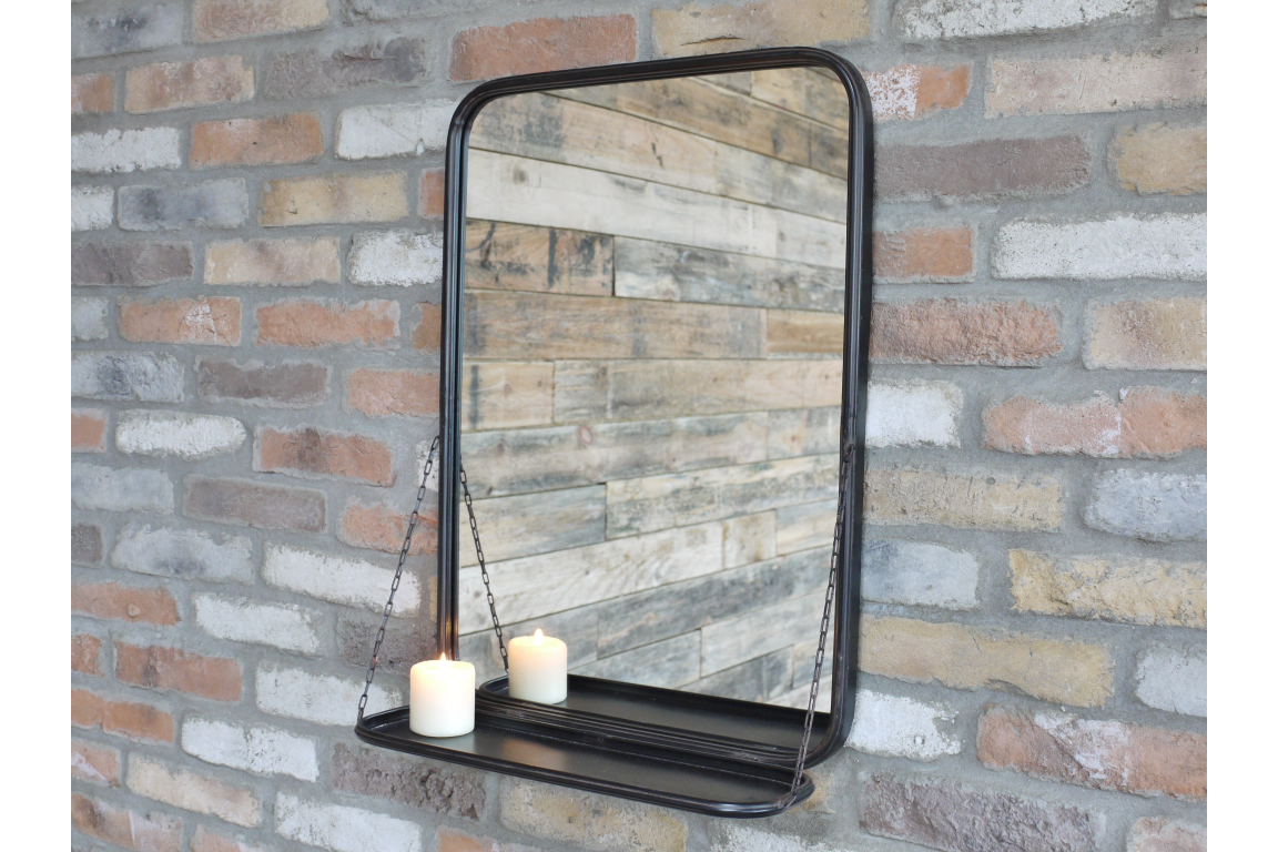 Rectangular black metal-framed wall mirror with attached shelf held by chains, shown against a brick wall.