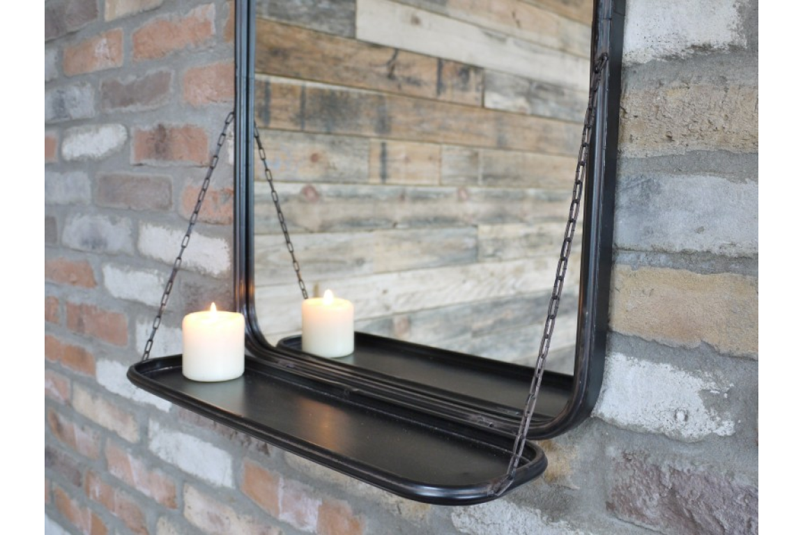 Black metal framed wall mirror with attached black shelf holding a lit white candle, mounted on brick wall.