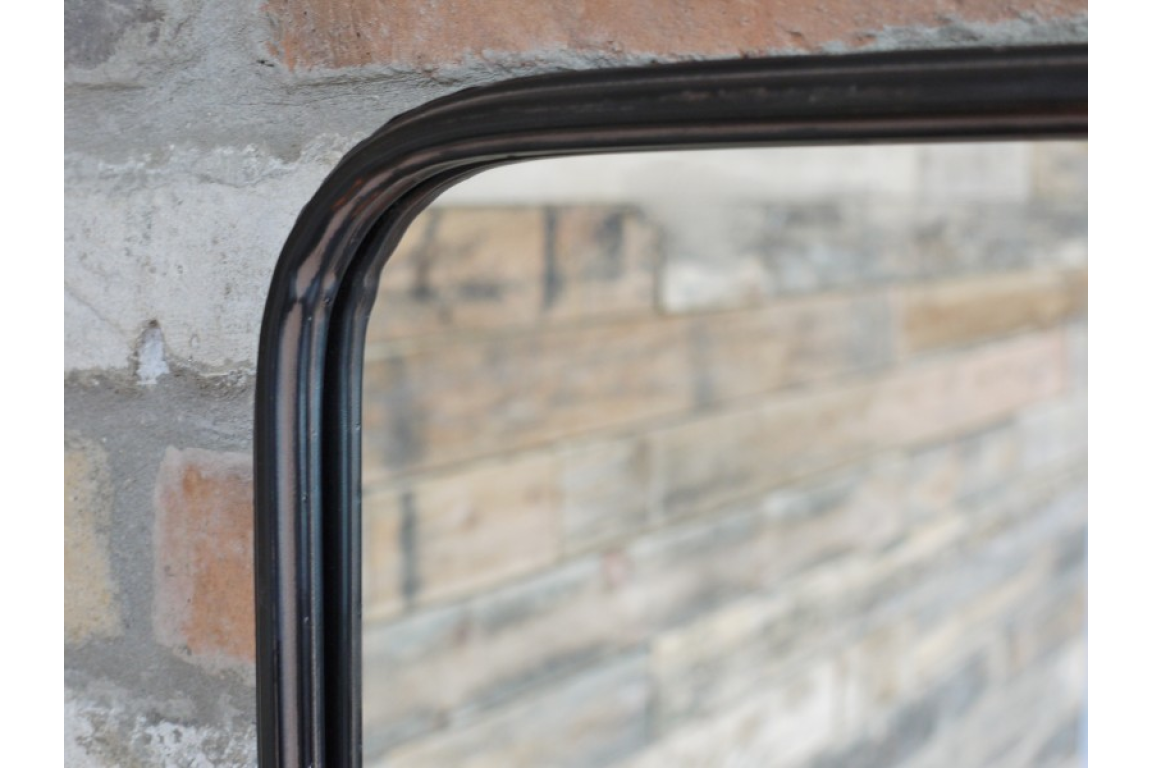 Black metal framed rectangular wall mirror with rounded corners and attached shelf, shown at side angle against brick wall.