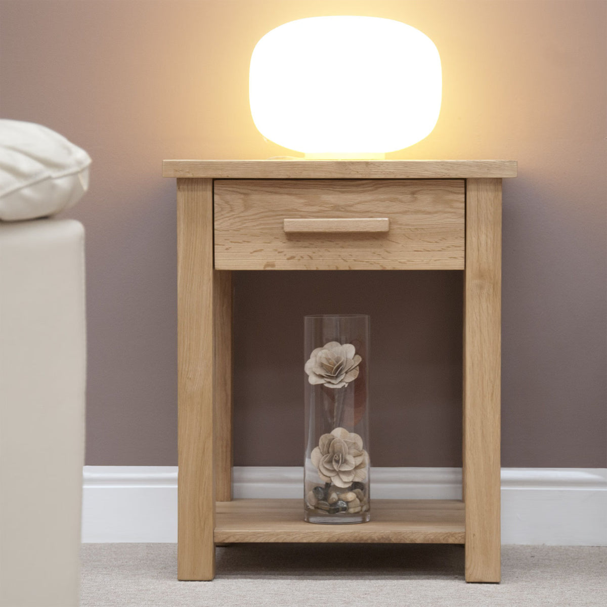 Oak bedside table with drawer, shelf, lamp and floral vase in modern bedroom