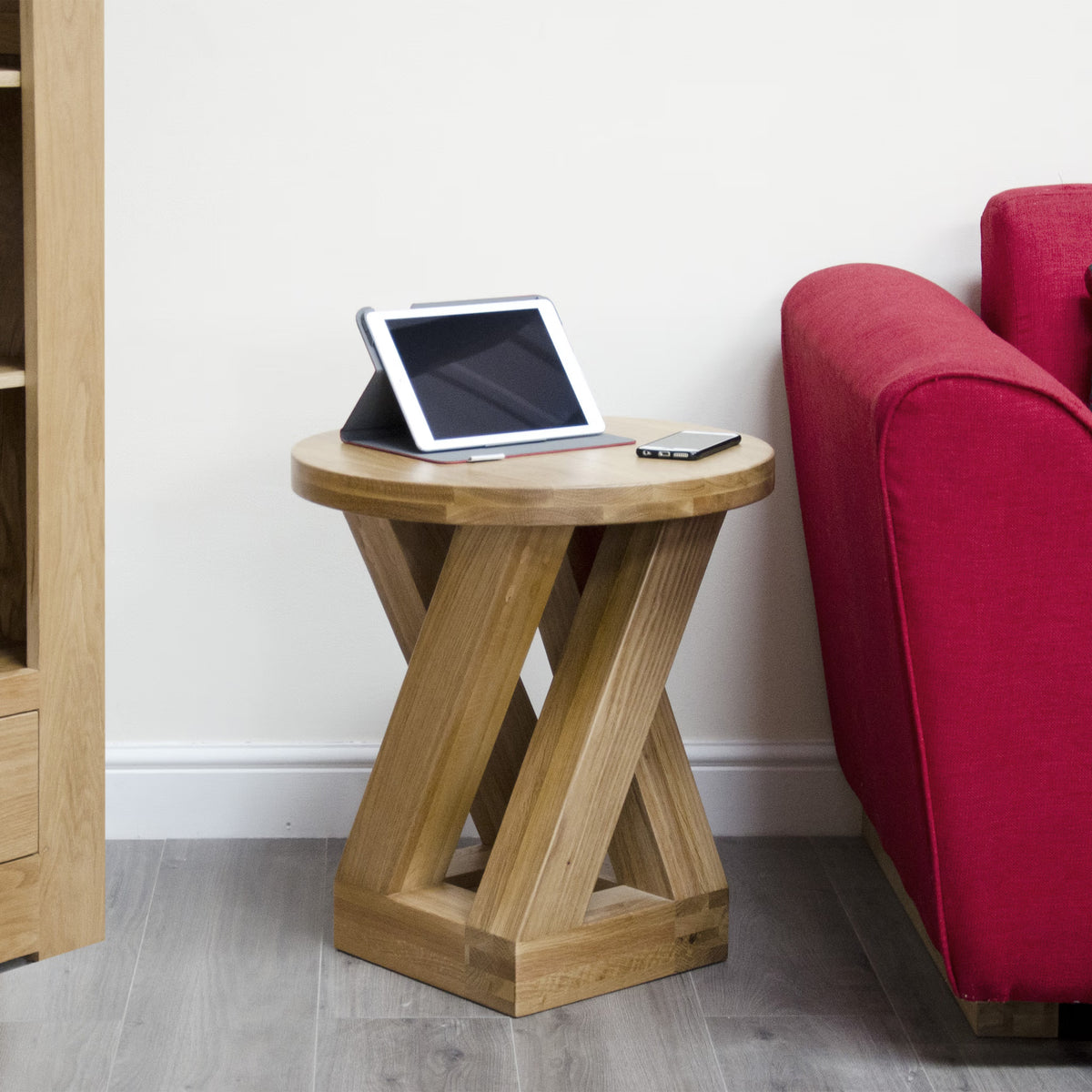 Oak side table with X-frame base beside red sofa, holding tablet and phone on top