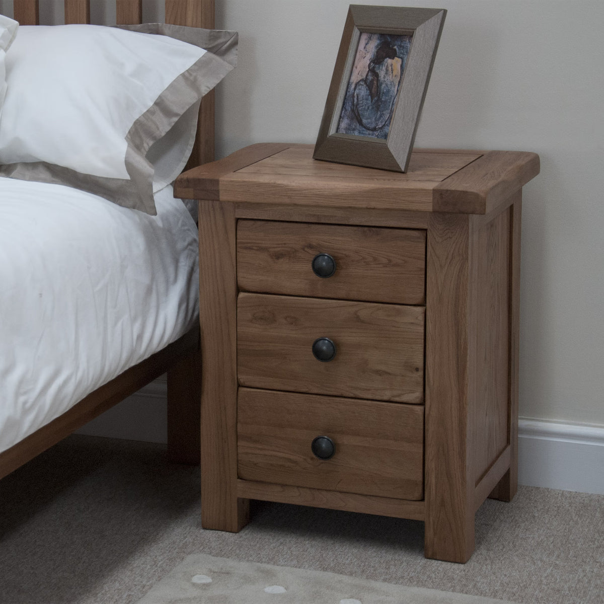 Oak bedside table with three drawers beside bed, framed photo on top